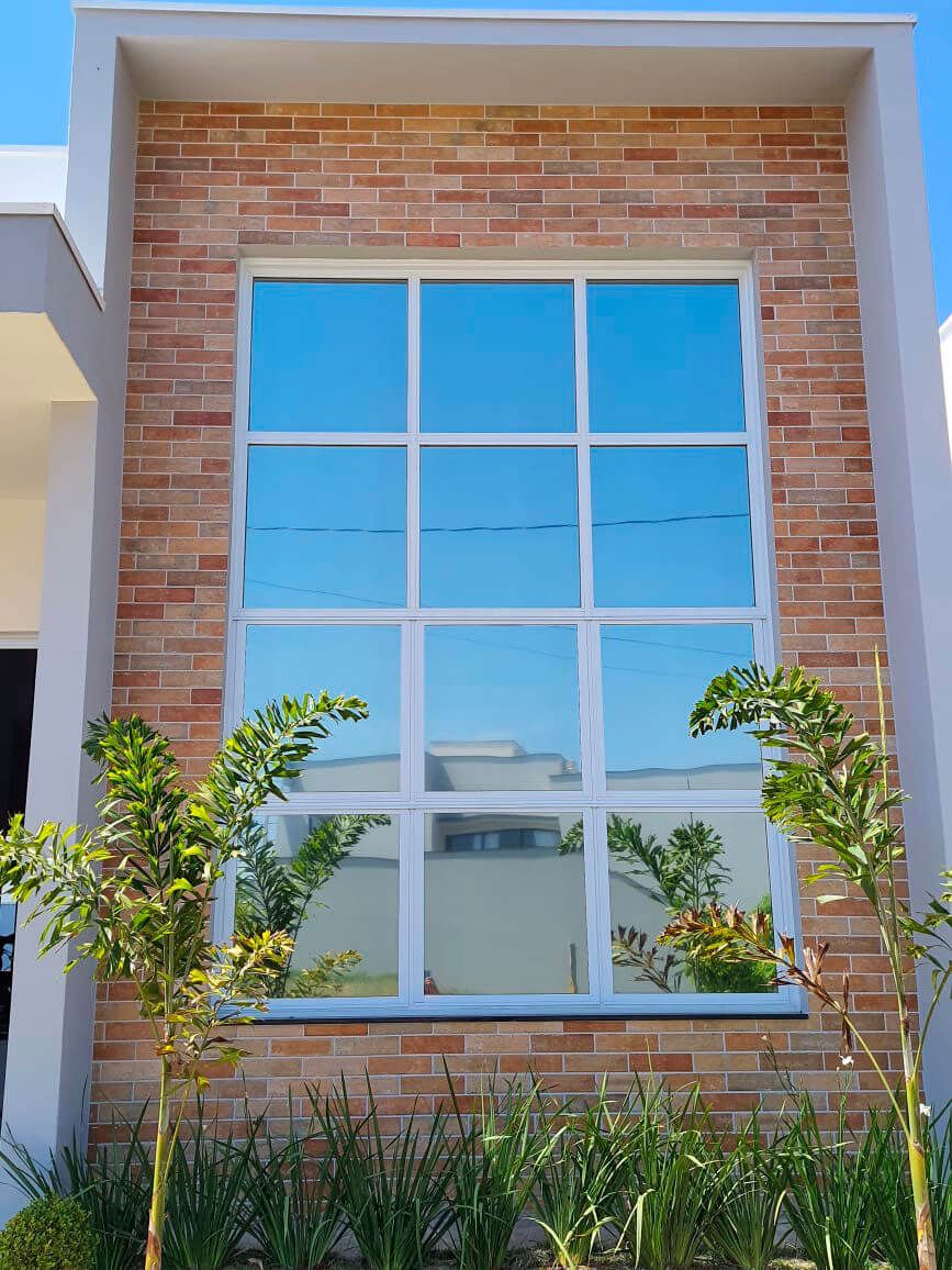 Janela residencial com película de controle solar refletindo o céu, em fachada de tijolo aparente